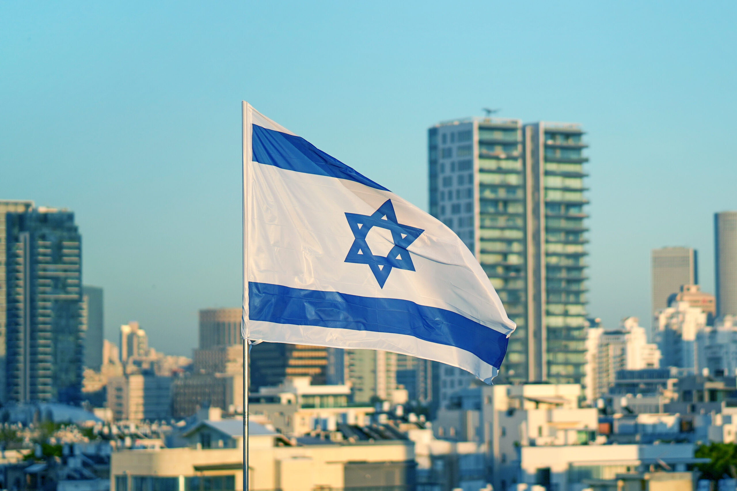 Flag of Israel on the background of modern city. Israeli flag against the background of skyscrapers in Tel Aviv in front of blue sky.